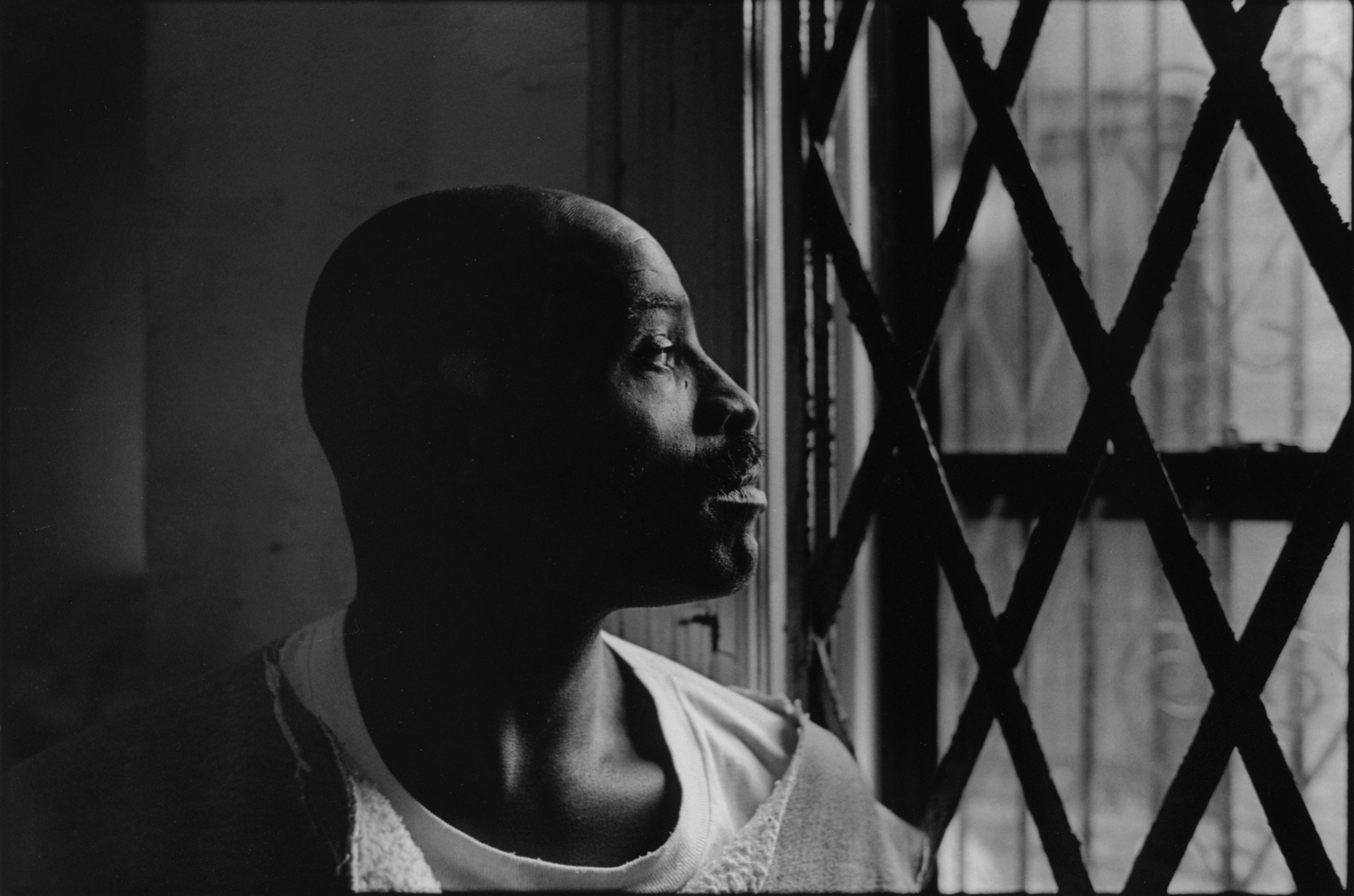 James looks out the front window of his apartment through the security grate on window. 1996. : Carmel Hill Harlem NYC 1995-2000 : BILL FOLEY 